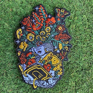 以下が含まれることがあります： 花柄の頭と本を持つ人物が描かれた刺繍パッチ。パッチは黒い背景に、赤、黄、オレンジの色合いのカラフルな花が描かれています。人物の顔は水色で、本は金色の縁取りがされています。