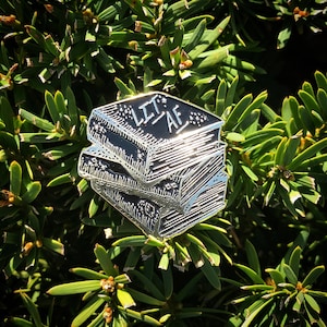 Op de afbeelding: Een zilverkleurige emaille pin met een stapel boeken met de tekst "LIT AF" op het bovenste boek.