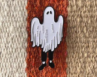 Puede incluir: Pin de esmalte de fantasma blanco con piernas y brazos negros. El pin está sobre un fondo texturizado naranja y marrón.
