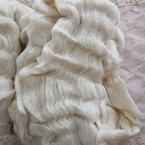 May include: Close-up of off-white yarn, tightly bundled and textured. The yarn appears soft and fluffy, with visible strands. The background is a light pink fabric with a delicate lace pattern, suggesting a craft or textile theme.