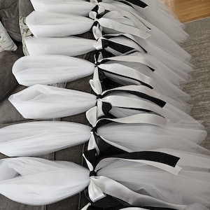 May include: Several white tulle skirts with black and white satin ribbon bows are arranged on a gray sofa. The skirts are layered and appear fluffy. The black and white ribbon adds a contrasting detail to the otherwise all-white skirts.