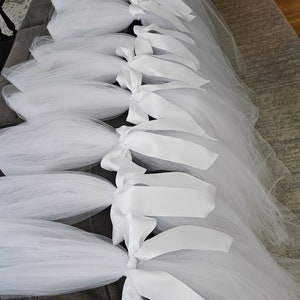 May include: Several white tulle skirts with satin ribbon bows are arranged on a gray surface. The skirts are bunched at the waist and tied with wide, white satin ribbons. The tulle is layered, creating a full, flowing appearance.
