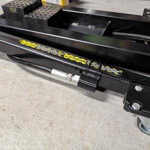 May include: Black hydraulic car lift with a warning label in yellow. The lift has a pair of rubber blocks and a set of wheels. The lift is designed to raise vehicles for maintenance and repair.