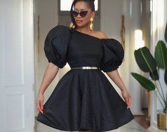 May include: A black dress with a one-shoulder neckline and puff sleeves. The dress has a fitted bodice and a flared skirt. The model is wearing a silver belt and black heels.