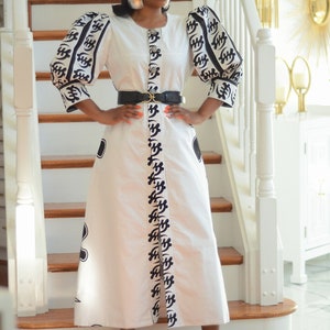Pode incluir: Um vestido maxi branco com mangas de padrão preto e branco e um cinto preto. O vestido tem uma faixa de padrão preto e branco na frente. A modelo está usando óculos de sol pretos e grandes brincos de prata.