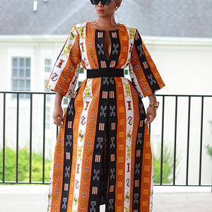 May include: A long, white, orange, and black dress with a geometric pattern. The dress has a deep V-neckline and a belt at the waist. The dress is worn with black pants and black shoes.