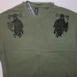 May include: A green t-shirt with a black graphic print of two armadillos hanging upside down. The armadillos are outlined in black and have a textured pattern.