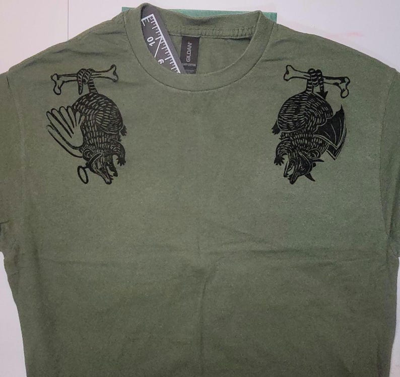 May include: A green t-shirt with a black graphic print of two armadillos hanging upside down. The armadillos are outlined in black and have a textured pattern.