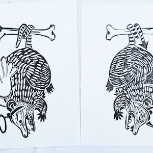May include: Two black and white linocut prints of an armadillo hanging upside down from a bone. The armadillo on the left has a wing and a magnifying glass. The armadillo on the right has a wing.