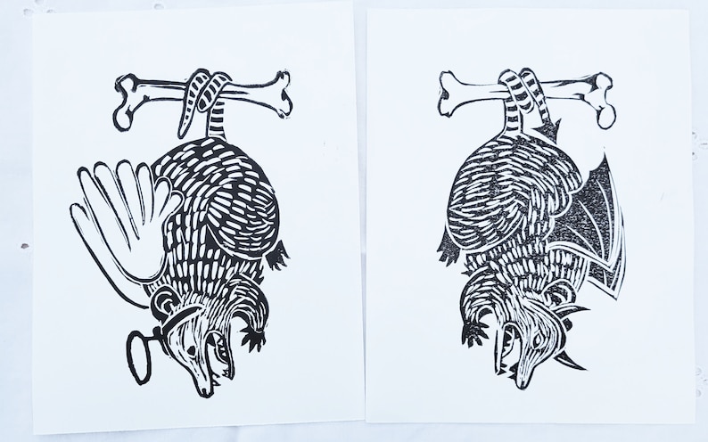 May include: Two black and white linocut prints of an armadillo hanging upside down from a bone. The armadillo on the left has a wing and a magnifying glass. The armadillo on the right has a wing.