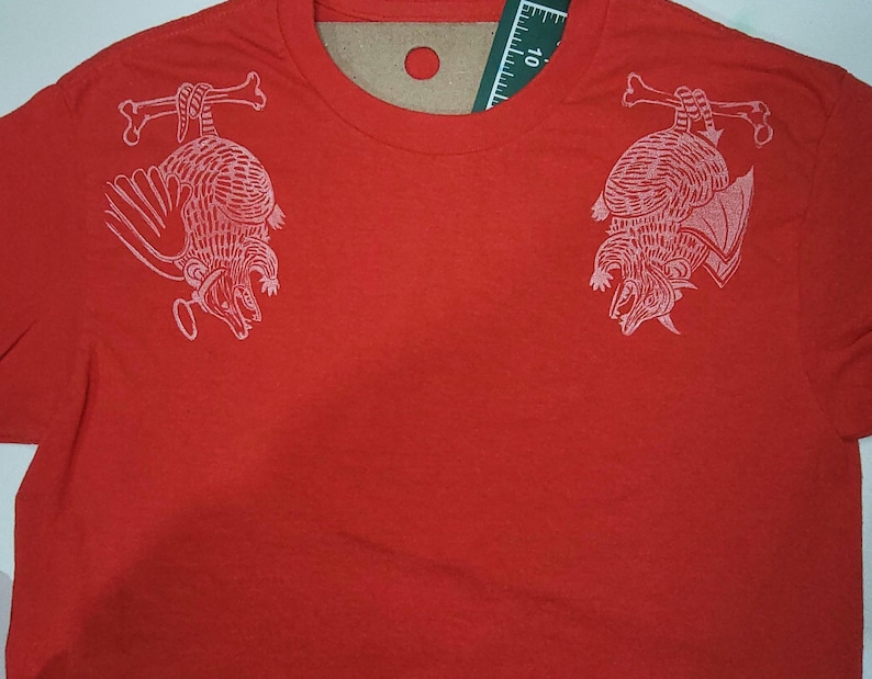 May include: Red t-shirt with a white graphic print of two armadillos hanging upside down from bones on each shoulder.