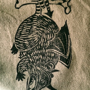 May include: A black and white illustration of a dragon holding two bones in its claws. The dragon is upside down and its wings are spread out. The illustration is printed on a light brown fabric.