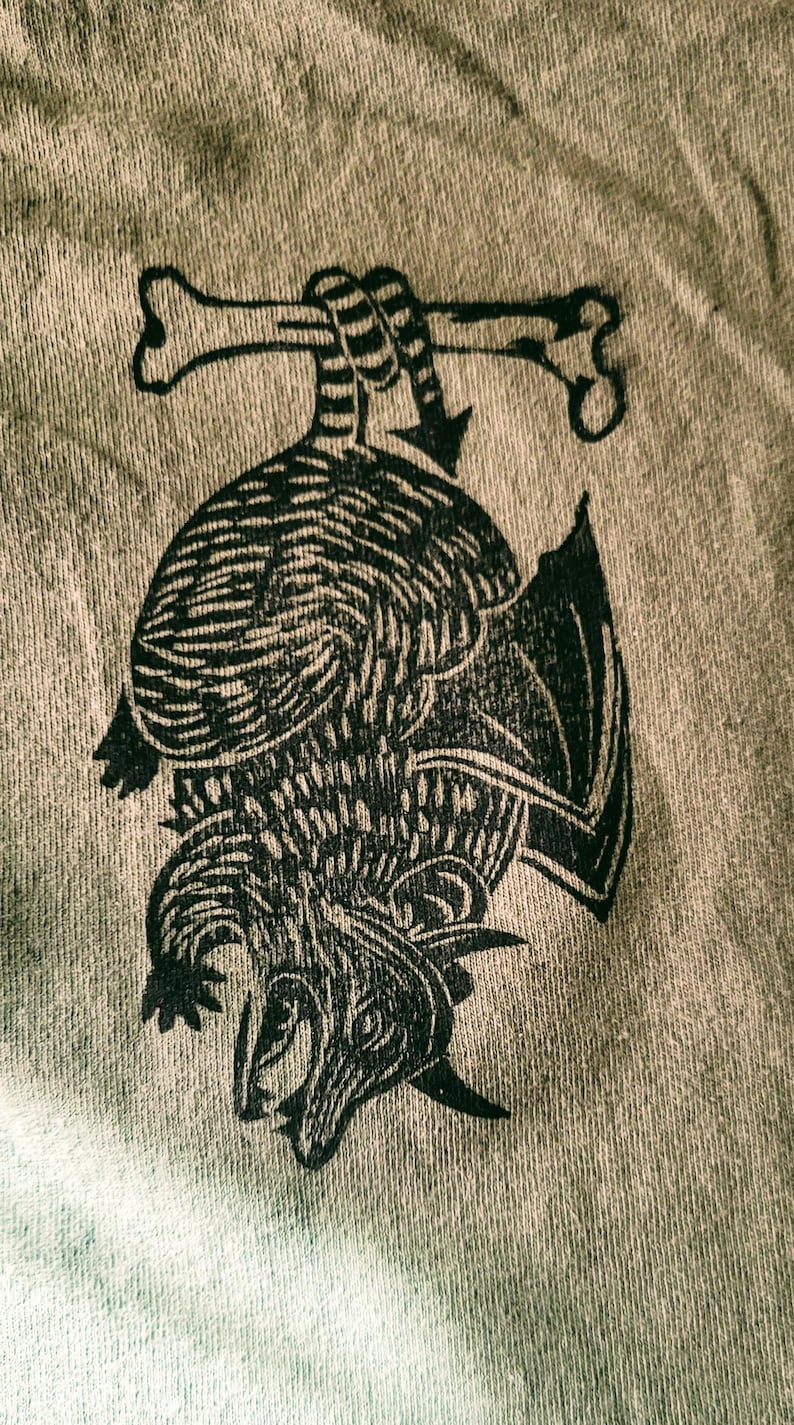 May include: A black and white illustration of a dragon holding two bones in its claws. The dragon is upside down and its wings are spread out. The illustration is printed on a light brown fabric.