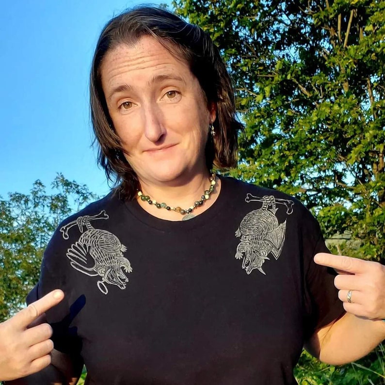 May include: A black t-shirt with a graphic print of two armadillos on the front. The armadillos are drawn in a stylized, almost skeletal, fashion. The person wearing the shirt is smiling and pointing at the shirt.