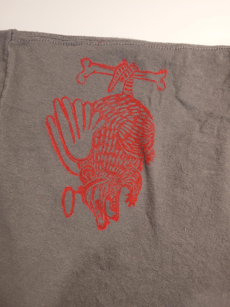 May include: A gray t-shirt with a red graphic of a possum hanging from two bones. The possum is drawn in a stylized, almost cartoon-like way.
