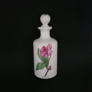 Może przedstawiać: Biała ceramiczna butelka z kwiatowym wzorem. Butelka ma korek i przedstawia różowy kwiat z zielonymi liśćmi. Tekst "Bauhinia monandra" jest nadrukowany na butelce.