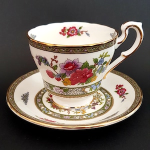 Puede incluir: Una taza de té y un platillo blancos con un borde dorado y un patrón floral. El patrón floral presenta flores rosas, rojas, moradas y amarillas con hojas verdes. La taza y el platillo tienen un diseño delicado e intrincado.