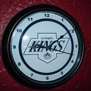 May include: Black and white clock with a Los Angeles Kings logo and the words "Los Angeles Kings" on the face. The clock has black hands and a black frame.