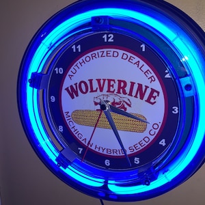 May include: A blue neon clock with a black face and white lettering. The clock face features a corn cob and a wolverine with the text "Wolverine Michigan Hybrid Seed Co. Authorized Dealer".