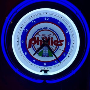 Peut inclure: Une horloge au néon blanche et bleue avec un logo des Phillies de Philadelphie. L'horloge a un anneau extérieur bleu et un anneau intérieur blanc avec des chiffres noirs.