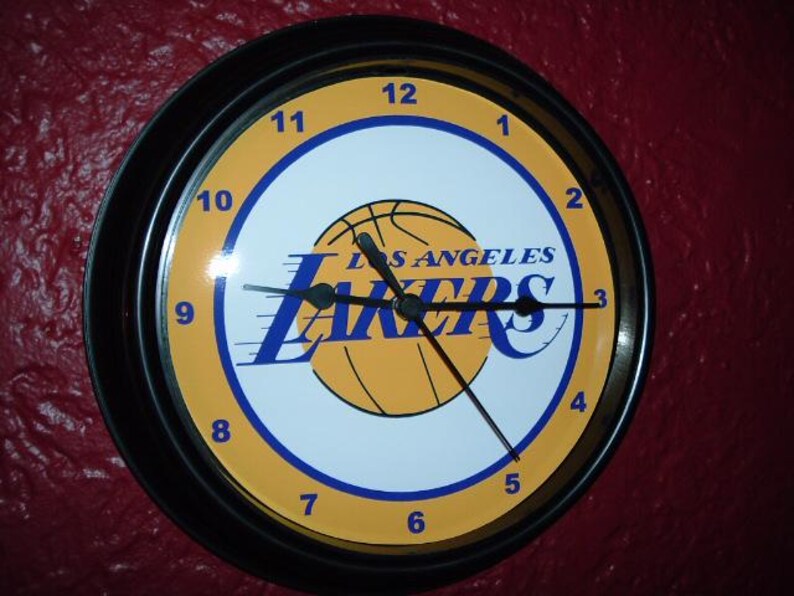 K&ouml;nnte beinhalten: Eine schwarz-gelbe Los Angeles Lakers Uhr mit einem Basketball-Motiv und dem Namen des Teams sowie der Stadt. Das Zifferblatt der Uhr hat die Zahlen 1 bis 12 und die Zeiger sind schwarz.