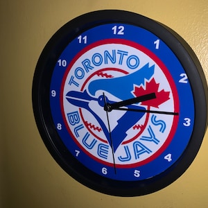 May include: A black and blue wall clock featuring the Toronto Blue Jays logo. The clock face displays the team's logo with the words "Toronto Blue Jays" and the numbers 1 to 12 around the edge.