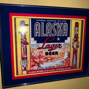 May include: A framed vintage advertisement for Alaska Lager Beer. The ad features a yellow background with red and blue borders. The text reads "Internal Revenue Tax Paid", "Alaska Lager Beer", "Contents 11 Fl. Ozs", "Permit WNU-1208", "Pure Glacial Water", and "Pilsener Brewing Co. of Alaska - Ketchikan, Alaska". The ad also features a cartoon image of a man in a red shirt and blue pants filling a bucket with water from a stream. There are two Native American totem poles on either side of the ad.