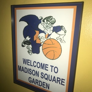 May include: A framed sign with the text "WELCOME TO MADISON SQUARE GARDEN." The sign features a cartoon character in blue and orange, holding a basketball. The sign has a blue border and is mounted on a yellow wall.