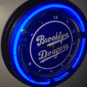 May include: A round, blue neon clock with the words "Brooklyn Dodgers" in white script. The clock has a black frame and a bright blue neon light around the perimeter. The clock hands are black.