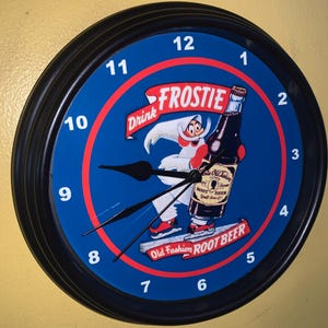 Puede incluir: Un reloj redondo negro y azul con un borde negro. La esfera del reloj presenta un personaje de dibujos animados sosteniendo una botella de cerveza de raíz con el texto "Drink Frostie Old Fashion Root Beer".