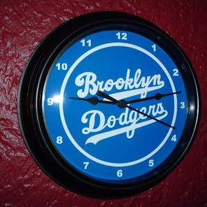 May include: A black and blue wall clock with white numbers and hands. The clock face features the words "Brooklyn Dodgers" in white lettering on a blue background.