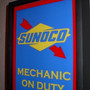 Puede incluir: Un letrero enmarcado en negro con un borde rojo y un fondo azul. El letrero tiene un logotipo amarillo y rojo con el texto "SUNOCO" y el texto "MECHANIC ON DUTY" en letras amarillas.