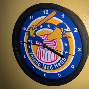 May include: A black and blue clock with a yellow, red, and white logo of a cartoon bird holding a baseball bat. The logo says "Toledo Mud Hens".