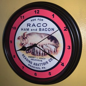 May include: Round wall clock with a black frame and a red and white face. The clock face features a pig with piglets and the text "RACO HAM and BACON". The clock also has the text "Reading Abattoir Co."