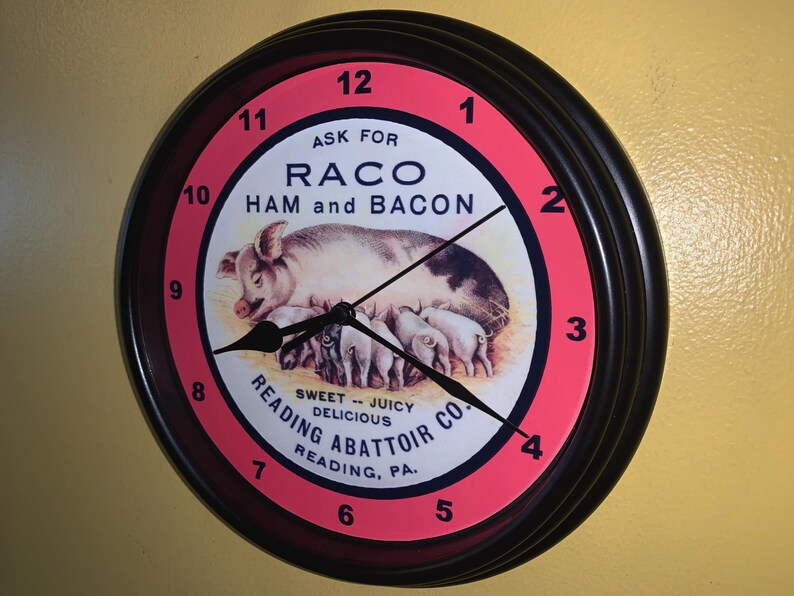 May include: Round wall clock with a black frame and a red and white face. The clock face features a pig with piglets and the text "RACO HAM and BACON". The clock also has the text "Reading Abattoir Co."