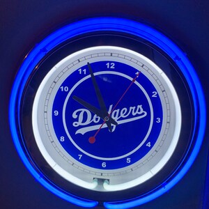 May include: A blue and white neon clock with the word "Dodgers" in a white script on a blue background. The clock has black hands and white numbers.