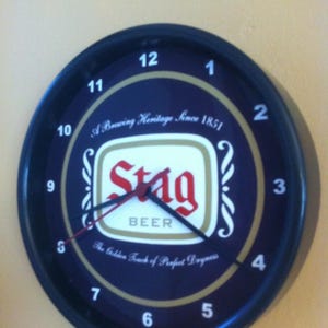 May include: A black and gold clock with a Stag Beer logo. The clock face is blue with white numbers and a red and black hour and minute hand. The logo says "A Brewing Heritage Since 1851" and "The Golden Touch of Perfect Dryness".