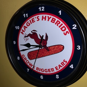 May include: A round wall clock with a black frame. The clock face features a red and white design with the words "HAGIE'S HYBRIDS" and "GROWS BIGGER EARS". A red cartoon rabbit jumps over an ear of corn.