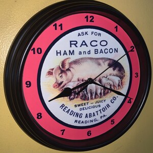 May include: A round wall clock with a black frame and a pink and white face. The clock face features a pig with piglets and the text "RACO HAM and BACON". The clock also has the text "READING ABATTOIR CO."