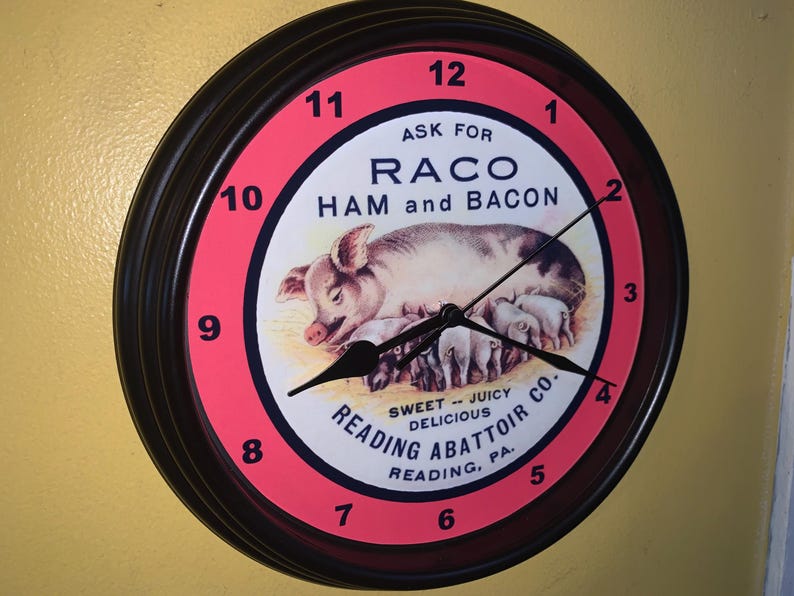 May include: A round wall clock with a black frame and a pink and white face. The clock face features a pig with piglets and the text "RACO HAM and BACON". The clock also has the text "READING ABATTOIR CO."