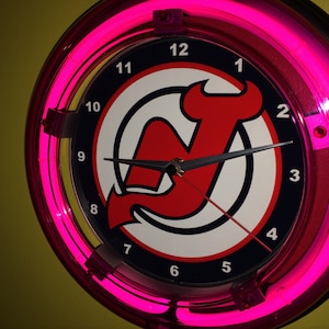 May include: A pink neon clock with a New Jersey Devils logo. The clock face has black numbers and a black and silver hand. The logo is red, white, and blue.
