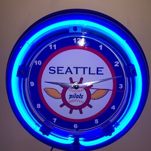 May include: A blue neon clock with a white face featuring the Seattle Pilots logo and the words "Seattle Pilots". The clock has a blue neon ring around the edge and black numbers.