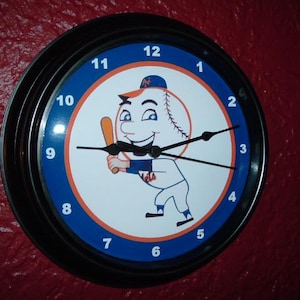 May include: Round wall clock with a black frame and a blue and orange face. The clock features a baseball character holding a bat. The numbers are in white, and the hands are black.