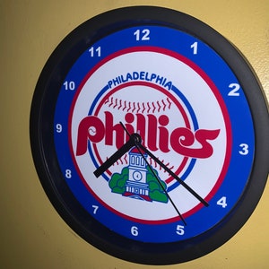 May include: A black and blue round wall clock with a white face featuring the Philadelphia Phillies logo. The logo includes a red and white baseball with the word "Phillies" in red letters. The clock face has black numbers and hands.