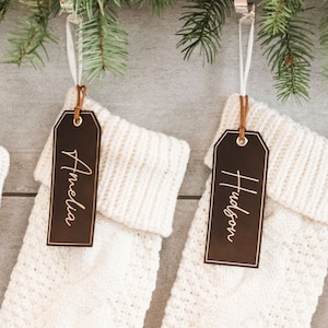 May include: Three white knitted stockings with brown leather tags hanging from a green garland. The tags are personalized with the names "Madison", "Amelia", and "Hudson".