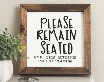 May include: A rustic wooden sign with the text "Please Remain Seated For The Entire Performance" in black lettering. The sign is sitting on a white toilet tank.