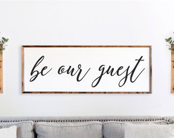 May include: A gray couch with three pillows, a white sign with the text "be our guest" hanging on the wall above the couch, and two wooden wall vases with greenery on either side of the sign.