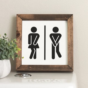 May include: A wooden framed sign with a white background and black stick figures depicting a woman and a man. The figures are in a stylized pose, suggesting they are using the restroom.
