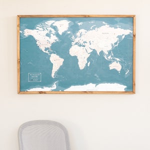 May include: A framed world map with a blue background and white continents. The map is decorated with small red and black pins. The map is hanging on a white wall above a desk with a laptop and a small plant.