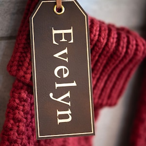 Op de afbeelding: Een rode gebreide sok met een bruin leren label waarop "Evelyn" in gouden letters staat.